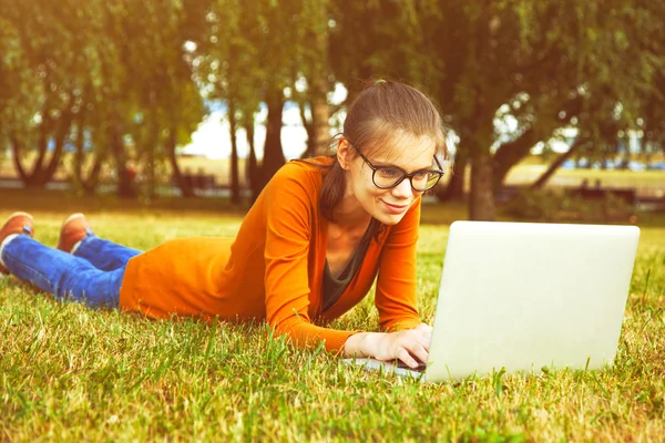 Смолящая девочка в очках, лежащая на траве и использующая ноутбук — стоковое фото
