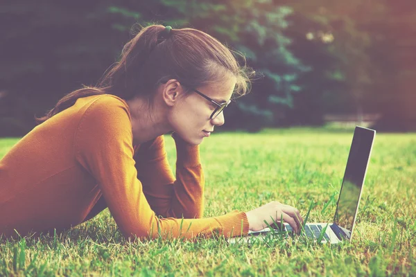 Leende tjej med glasögon ligga på gräset och använder laptop en — Stockfoto