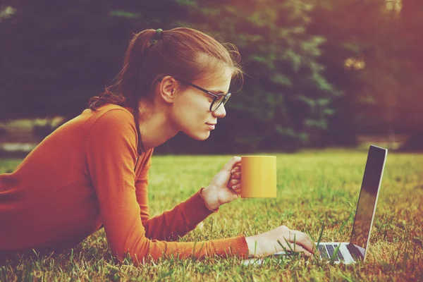 Gadis berbaring di rumput dan menggunakan laptop dan mengetik dengan kopi — Stok Foto