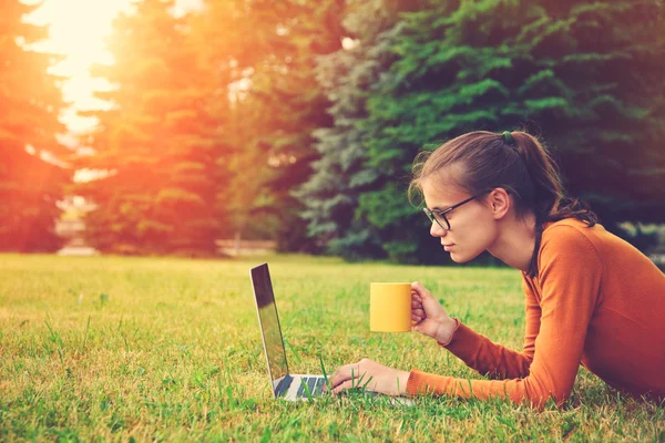 Chica acostada en la hierba y el uso de la computadora portátil y escribir con café — Foto de Stock