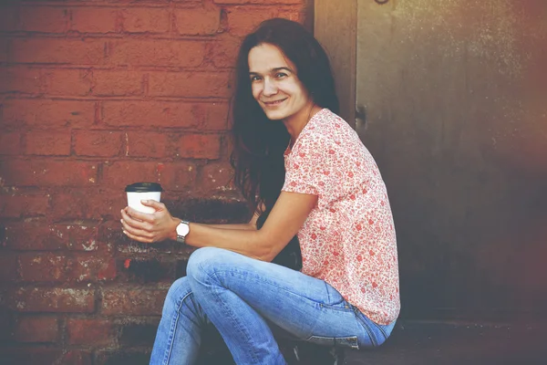 Χαμογελώντας όμορφη κοπέλα καθόταν κοντά σε τοίχο από τούβλα με τον πρωινό καφέ — Φωτογραφία Αρχείου