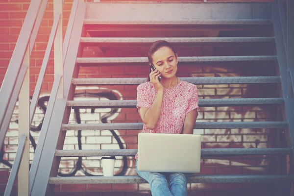 Gadis cantik dengan laptop berbicara telepon duduk di tangga tangga tangga — Stok Foto