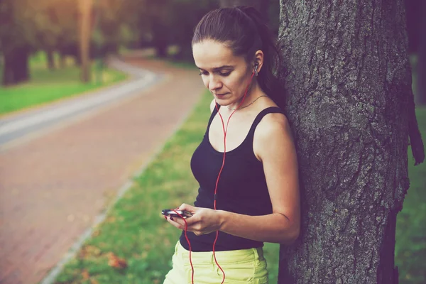 Mujer bastante sonriente corriendo en el parque con aplicación de teléfono inteligente y mus —  Fotos de Stock