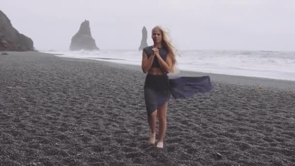 Modelo bonito que está ao lado da areia preta na praia em um dia ventoso — Vídeo de Stock