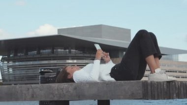 Beyaz kazaklı genç kız Portside Bench 'te uzanırken günün tadını çıkarıyor.