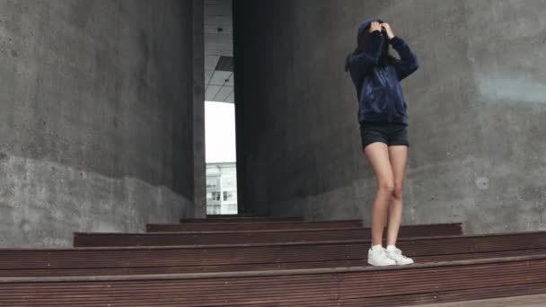 Teenage Model Standing by Stairs with Walls in Between Putting on Hoodie Jacket — Stock Video