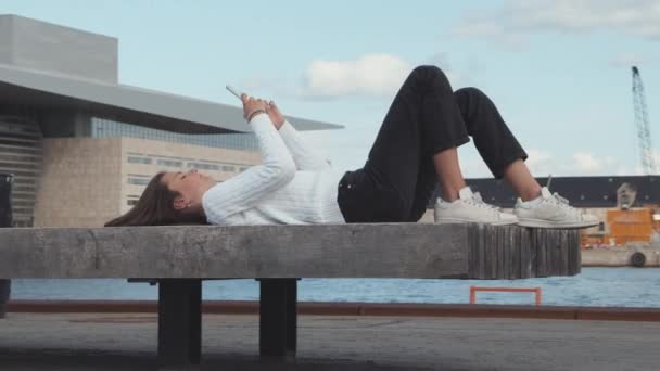 Teenager mit weißem Pullover legt sich auf Backbordbank gegen blauen Himmel — Stockvideo