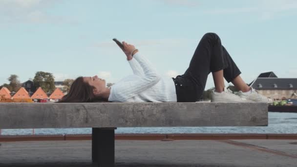 Gadis remaja dengan Sweater Putih Berbaring Bawah oleh Dockside Bench Menggunakan ponselnya — Stok Video