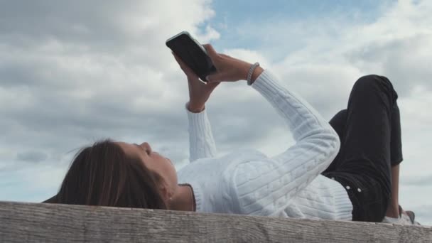 Adolescente en el puerto de Copenhague acostada en un banco al aire libre usando su teléfono — Vídeos de Stock