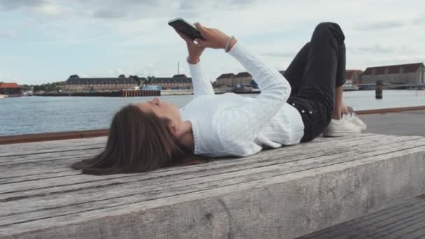 Tonåring Använda sin telefon på bänk liggandes vid Köpenhamns Dockside — Stockvideo