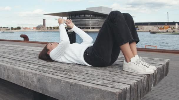 Frumoasă adolescentă care-și folosește telefonul zâmbind în timp ce stă întinsă pe docuri în Danemarca — Videoclip de stoc