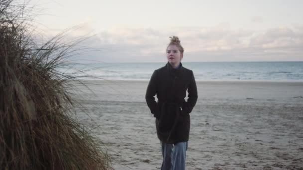 Roodharige meisje in zwart jas lopen op het strand — Stockvideo