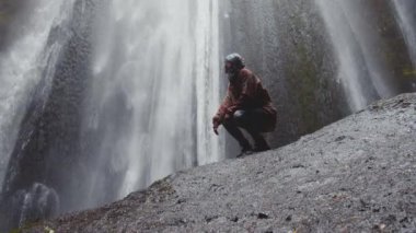 Şelalenin yanındaki kayada çömelmiş bir kadın