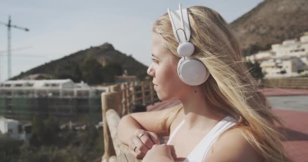 Lady en auriculares blancos inclinados sobre bordas de madera para admirar la vista de la montaña — Vídeo de stock