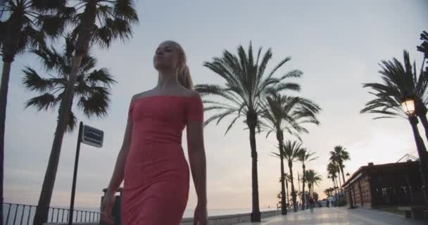 Schöne Frau in rosa Kleid sieht gerade aus und geht auf der Strandpromenade — Stockvideo