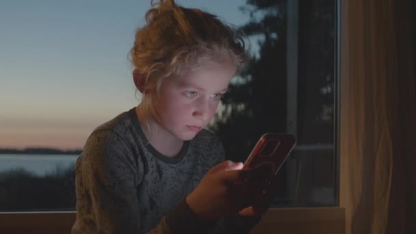 Niño iluminado por Smartphone por la ventana al anochecer — Vídeo de stock