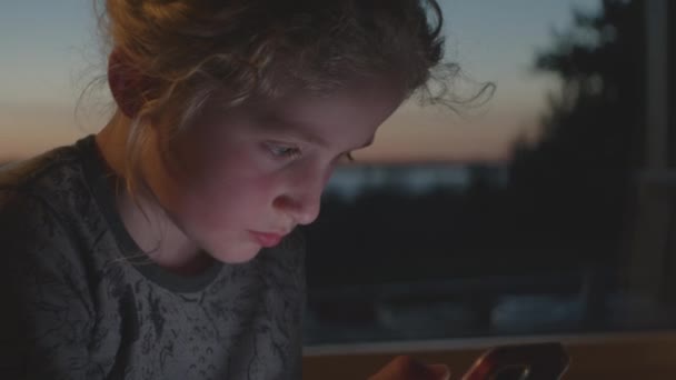 Niño jugando en el teléfono inteligente por la ventana al anochecer — Vídeo de stock