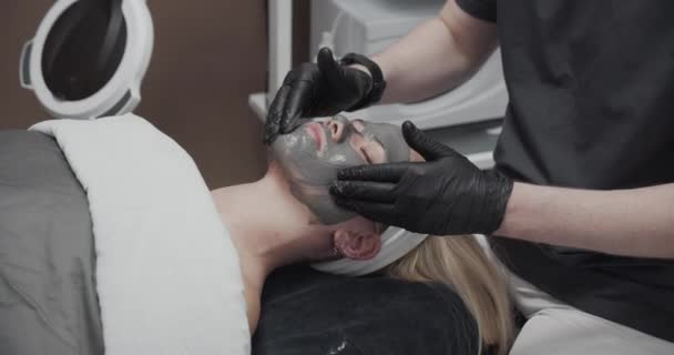 Kosmetikerin massiert graue Gesichtsmaske auf dem Gesicht einer Frau — Stockvideo