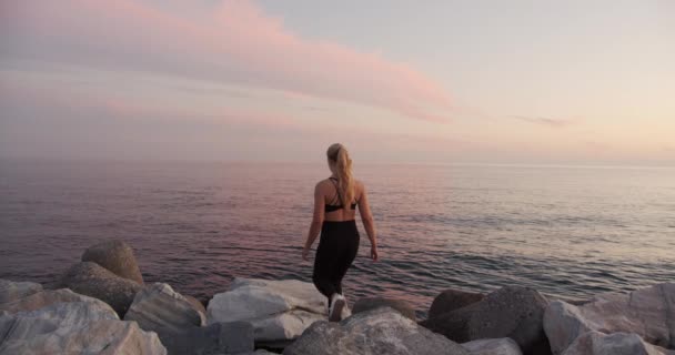 Ung idrottsman tittar på solnedgång över havet från stenar — Stockvideo