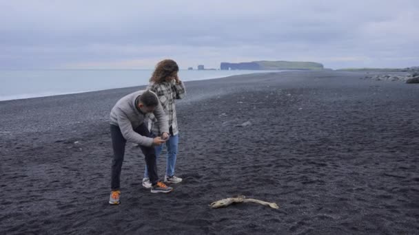 Piękna para przechwytująca zdjęcia szkieletu ryby na Reynisfjara Black Sand — Wideo stockowe