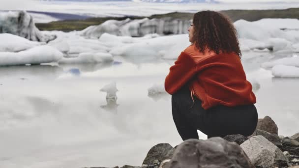 Uitzicht op het bevroren meer en smeltende ijsberg en een vrouw die bij de rotsen zit — Stockvideo