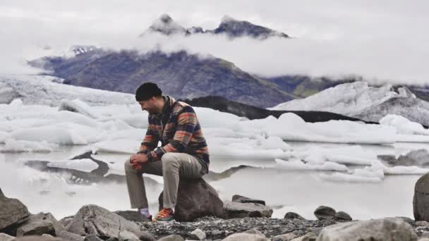 Muž turista sedí na skále, zatímco pózuje pro fotografii s ledovcem na pozadí — Stock video