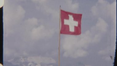 Bir kişi, İsviçre bayrağının yanında dikilirken, bir direğin üzerinde, karlı dağlardan oluşan bir zemin üzerinde sallanıyor, 8 mm 'lik bir filme çekiliyor, sakin ve görkemli bir görsel hava yaratıyor..