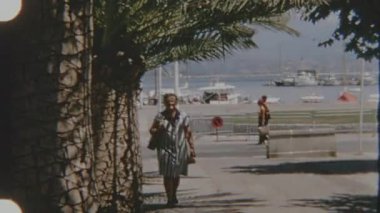 Bu 1970 'lerin 8mm' lik film görüntülerinde Korsika 'da güzel bir sahil gezisi görülüyor. Çizgili elbiseli bir kadın, uzun palmiye ağaçlarının altında manzaralı bir arka plan ve sakin bir limana doğru yürüyor.