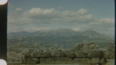 8mm 'lik bir filmde çekilen bu video, bulutlarla benekli mavi bir gökyüzünün altındaki sakin bir dağ zeminine karşı antik taş kalıntılarını sergiliyor..