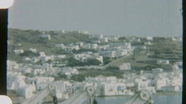 Bir geminin güvertesinden Mykonos 'un görüntüsü, 8 mm' lik bir film, bir yamaçtaki beyaz binaları çekiyor, cankurtaran botları ve ön plandaki sakin denizle.