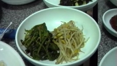 Büyük bir kase dolusu baharatlı ıspanak, fasulye filizi ve taze sebzeli kimchi yemek masasında birkaç küçük kase Kore mezesi ile çevrilidir.