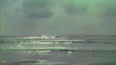 Geniş, yansıtıcı bir kıyı şeridi alçak gelgitte sisli bir gökyüzünün altında uzanır nazik dalgalar süzülür ve suların kenarında uzak siluetli bir figür durur.