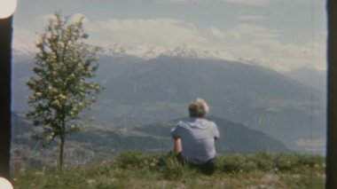 Bir adam, karla kaplı dağlara tepeden bakan çimenli bir tepede oturur ve gündüz vakti çekilen 8 mm 'lik bir filme çekilir..