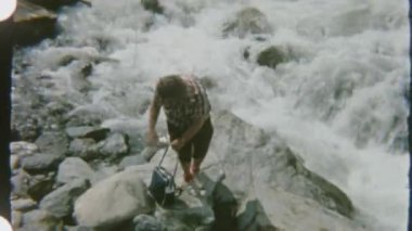 Ekose gömlek ve güneş gözlüğü takan bir kadın, kayalık bir nehrin kenarında, akıntılı bir nehirde duruyor. Gündüz vakti çekilen 8 mm 'lik filmle..