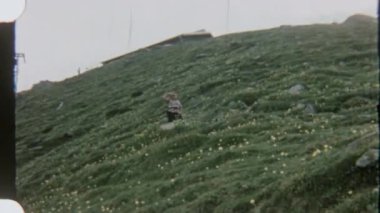 Dişi bir yürüyüşçü, bulutlu bir gökyüzünün altındaki karlı dağlara doğru çimli bir tepeye tırmanır ve gündüz vakti çekilen 8 mm 'lik filmle yakalanır..