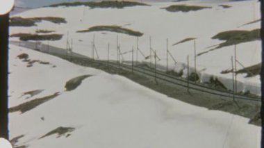 Bir tren karlı bir dağ yamacında ilerliyor, 8 mm 'lik filme çekiliyor, sakin ve nostaljik bir görsel hava yaratıyor. Ön vagonda iki kişi görünüyor..