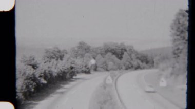 Siyah ve beyaz 8 mm 'lik kameralar, verimli ağaçlar ve sakin doğal manzarayla çevrili virajlı bir yolda pürüzsüz giden araçları yakalıyor.