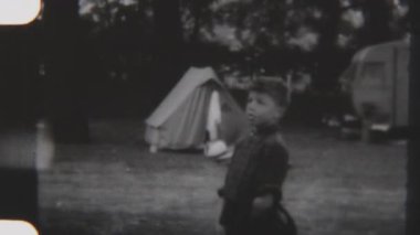 Genç bir çocuk ormanlık bir kamp alanında duruyor, çadırı ve karavanı yemyeşil doğayla çevrili, siyah ve beyazla çevrili.
