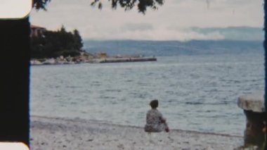 Plajda huzur içinde oturup kıyıya vuran canlı mavi okyanus dalgalarını seyreden bir kadının nostaljik ve huzurlu bir sahili yakaladığı 8mm 'lik bir görüntü.