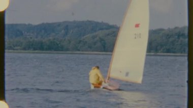Bir adam sakin bir su kütlesi boyunca yelken açar, küçük tekneyi dengelemek için mücadele eder, rüzgar ana yelkeni yakalar, gündüz vakti çekilen 8 mm 'lik filmle..
