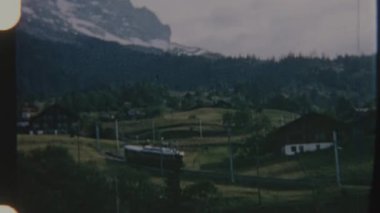 Bir tren, dağlarda yuva yapmış, 8 mm 'lik bir filme çekilen pitoresk bir köyden geçiyor. Sahnede geleneksel evler ve yemyeşil alanlar sergileniyor. Huzurlu ve nostaljik bir görsel ortam yaratıyor..