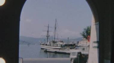 Büyük bir yelkenli ve arka planda dağlarla birlikte geçen motorlu araçların yer aldığı, taştan kemerli bir kemer yolundan izlenen bir limanın 8 mm 'lik Retro filmi. Sahne huzurlu ve nostaljik bir ruh hali yakalıyor..