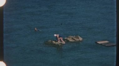 Bir kadın sakin deniz sularında bir kayanın üzerinde oturuyor, yanında başka biri yüzüyor, nostaljik 8 mm 'lik bir filme çekiliyor..