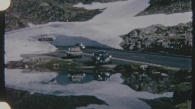8mm 'lik bir filme çekilen eski model bir araba, dağ yolunda karlı bir manzaraya karşı park edilmiş, nostaljik ve manzaralı bir görsel hava yaratıyor..