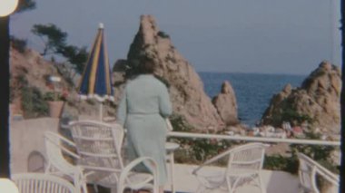Açık yeşil elbiseli bir kadın, denize bakan bir balkonda beyaz bir sandalyede oturuyor. Gündüz vakti çekilen 8 mm 'lik filmle çekiliyor..