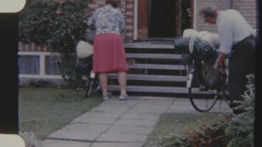 Ön bahçelerinde bisiklet gezisi öncesi bisiklet çantalarını ayarlayan bir çiftin klasik filmi, arka planda görünen tuğla cephe.