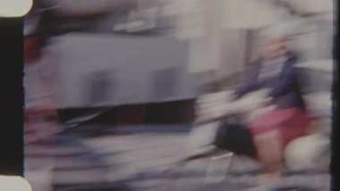 Bir kadın, 8 mm 'lik bir filme çekilen bir feribota çantalarla dolu bir bisiklete biniyor. Sahne bir seyahat ve hareket hissi veriyor..