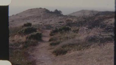 Bir kadın çimenli bir tepede pis bir patika boyunca yürüyor, gündüz vakti 8 mm 'lik bir filme çekiliyor. Sahne huzurlu ve huzurlu bir görsel hava veriyor..