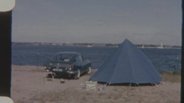 Kumlu bir sahilde mavi bir çadırın yanına park edilmiş klasik bir araba, gündüz vakti 8 mm 'lik bir filme çekiliyor, sakin ve sakin bir ruh hali uyandırıyor..