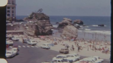 Canlı bir 8 mm 'lik film, insanların güneşlendiği ve yüzdüğü, yakınlarda park edilmiş arabaların, kıyıya vuran dalgaların ve arka planda çarpışan kaya oluşumlarının yer aldığı canlı bir plaj sahnesi çeker..